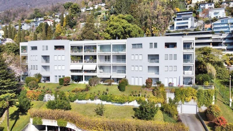 Modern apartment building, white exterior, multiple balconies, surrounded by greenery