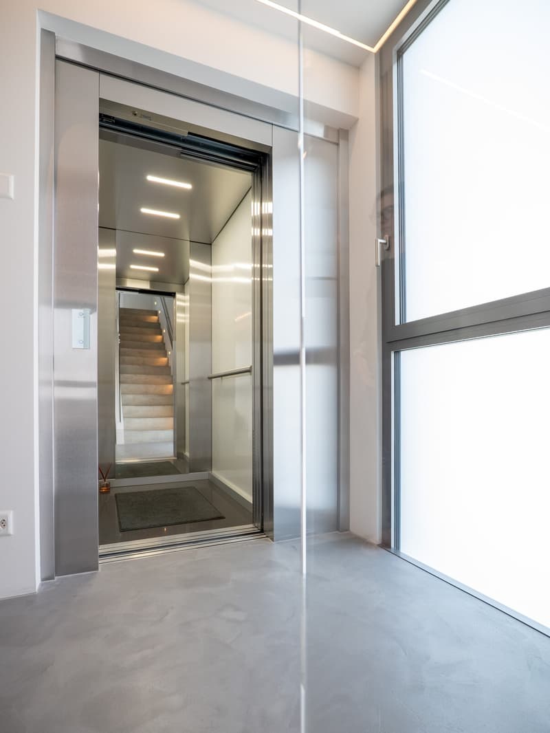 Modern elevator with glass doors, stainless steel interior, illuminated by ceiling lights, open to a staircase leading to upper floors, concrete floor, large window