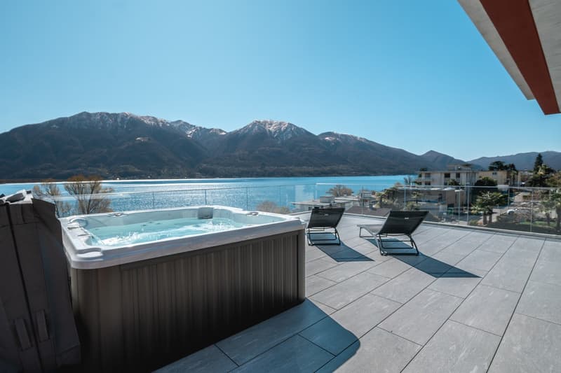 Outdoor terrace with a hot tub, two lounge chairs, and a glass railing overlooking a lake and mountains.