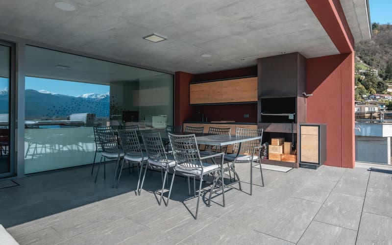 Outdoor dining area with table and chairs, large glass wall, wooden cabinets, storage boxes, outdoor grill, tiled floor, mountain view