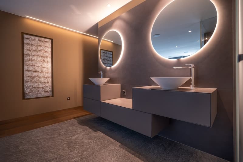 modern bathroom with double sink, round mirrors, wooden floor, gray walls, artwork