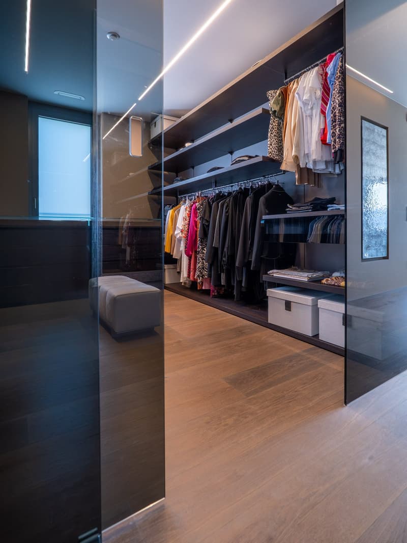 walk-in closet with glass walls, wooden floor, built-in shelves, hanging clothes, storage boxes, ottoman
