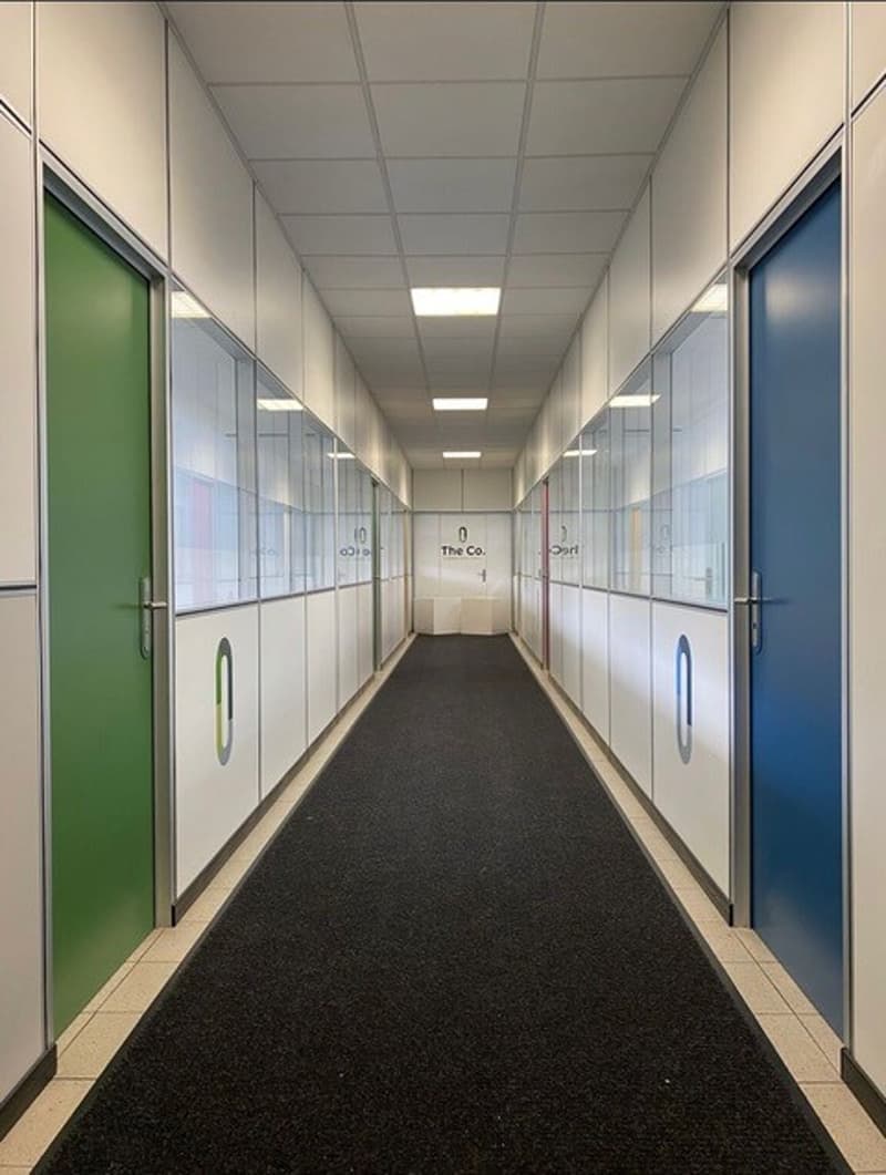 The image shows a long, narrow hallway with a carpeted floor and a suspended ceiling. The walls are lined with glass-paneled doors, some of which are green and others are blue. The hallway appears to be in a commercial or office building setting.