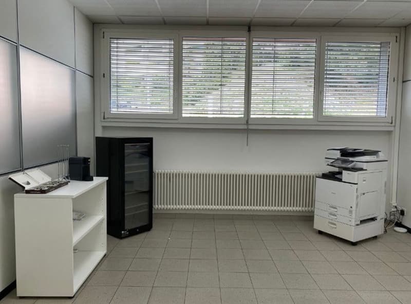 The image shows an office space with large windows, a desk, a printer/copier, and a computer tower. The room has a minimalist, clean design with white walls and tile flooring.
