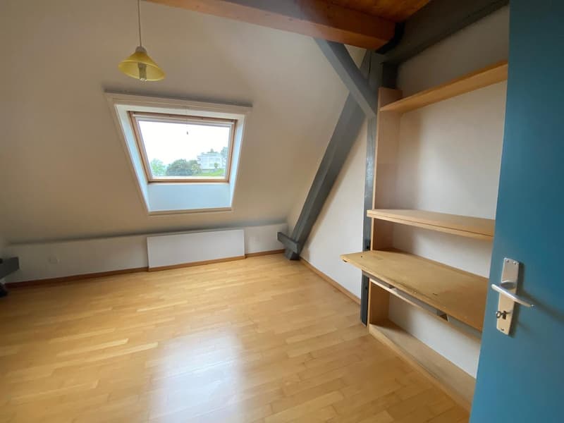 Attic bedroom with hardwood floors, white walls, wooden beams, and a skylight window providing natural light. The room features built-in shelving and storage space.