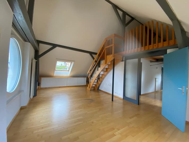 Spacious loft-style room with high ceilings, wooden staircase leading to a mezzanine level, hardwood flooring, and a skylight window providing natural light.