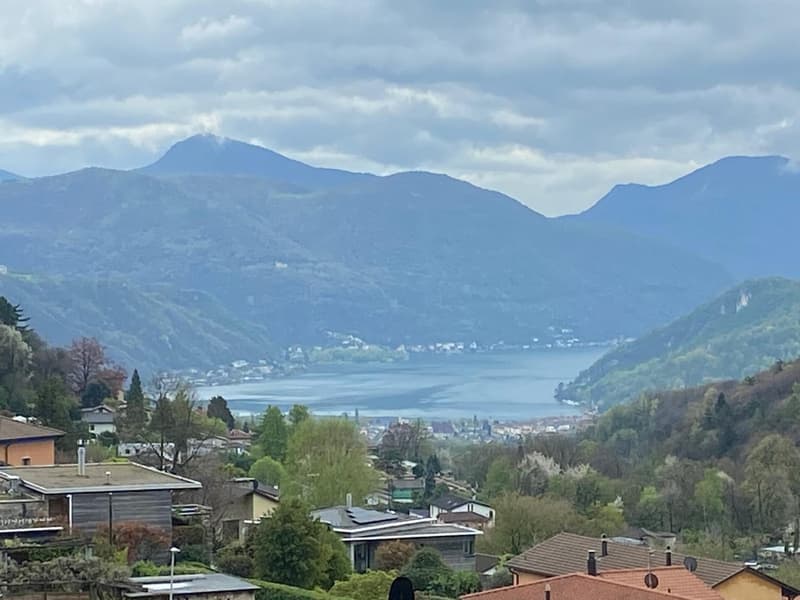 The image shows a scenic view of a mountainous landscape with a lake in the foreground. The town is nestled between the mountains, with houses and buildings visible. The sky is overcast with clouds, creating a moody and atmospheric setting.