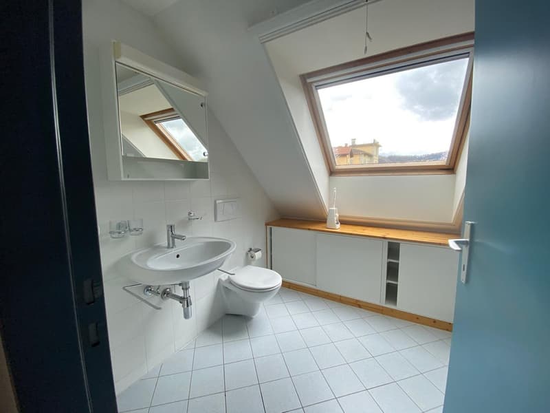 Bathroom with white tiles, a sink, a toilet, and a window with a view of the outdoors.