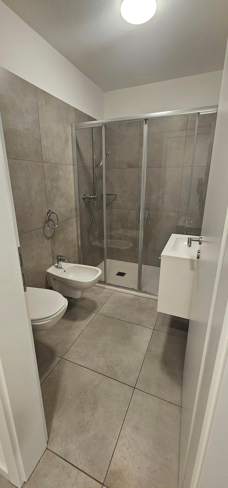 modern and spacious bathroom with white sink, separate bidet, glass walk-in shower, gray tile flooring