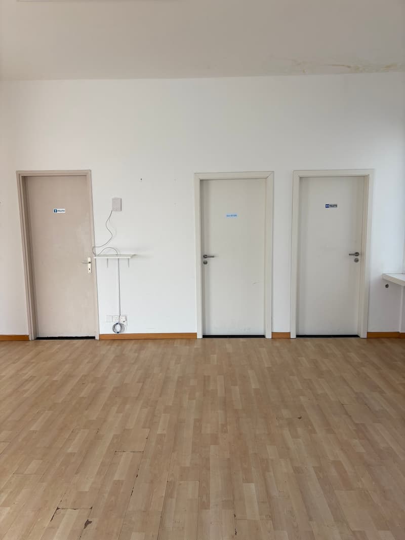 Empty room, white walls, wooden floor, three doors, power outlets, bathroom sign