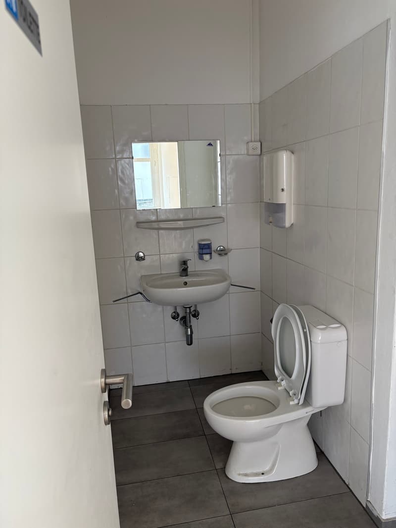 Compact bathroom with white tiled walls, a toilet, a sink, a soap dispenser, and a small mirror.