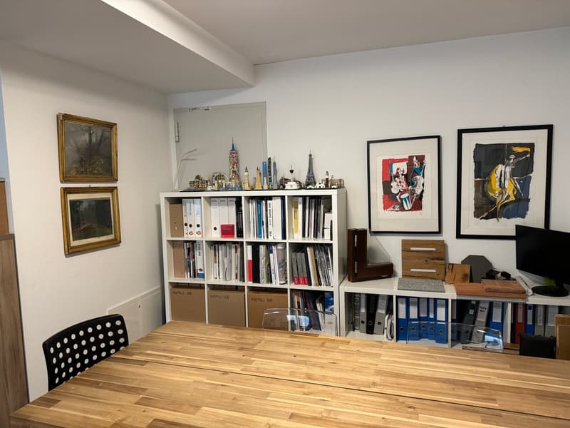 a room with a wooden table, two chairs, a bookshelf with many books, two framed pictures on the wall, and a monitor