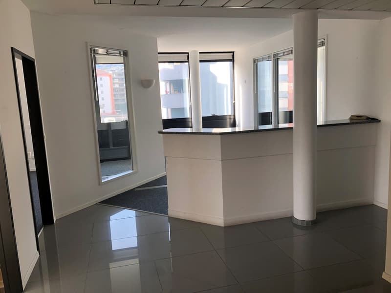 An empty room with white walls, gray tiled floor, a white reception desk, a mirror, and a wall-mounted lamp. There are large windows with black frames and a black carpet on the floor.