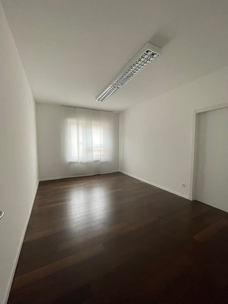 Empty room with wooden floors, white walls, and white curtains. One window with blinds and power outlet. A ceiling light.