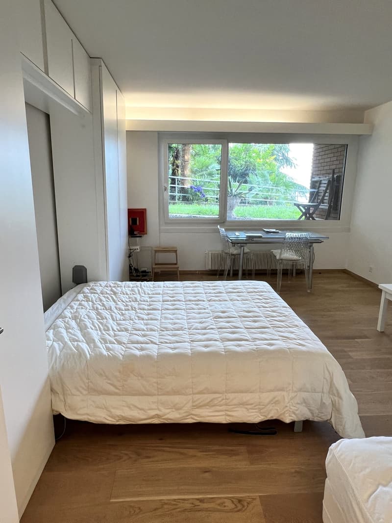 Single bed, hardwood floor, white walls, large window, bedside table, white cabinets, desk and chairs in front of the window