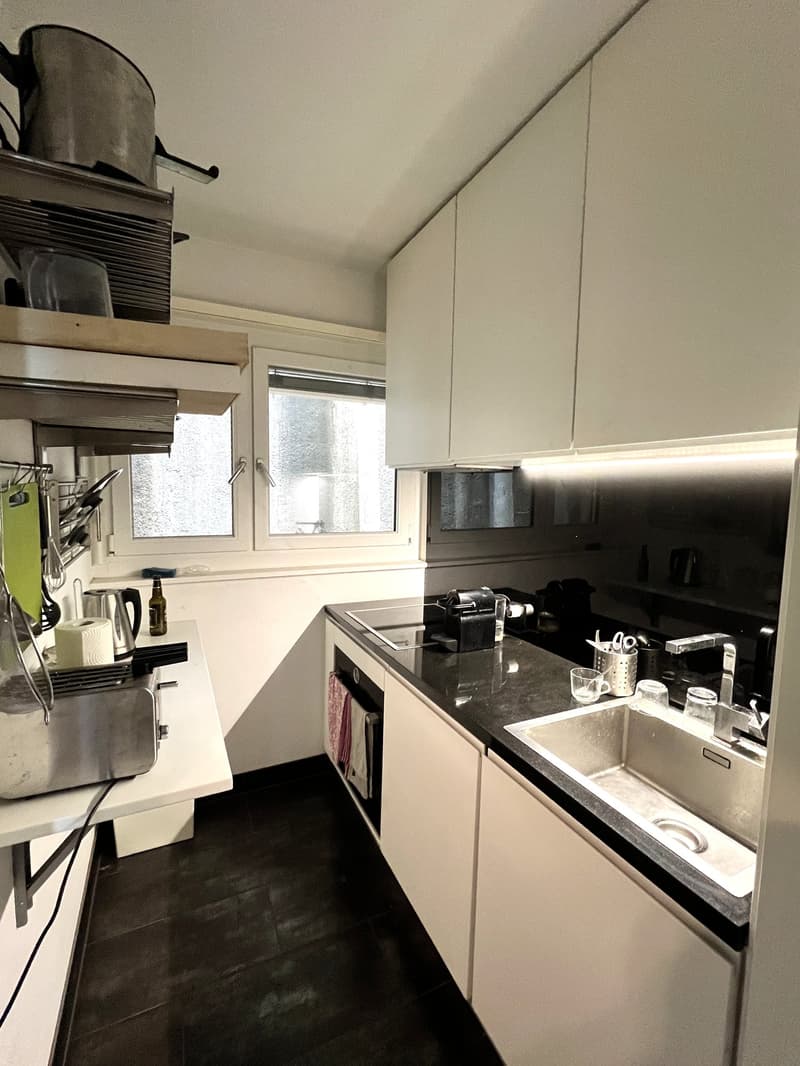 Modern kitchen with white cabinets, black countertops, stainless steel sink and appliances. Natural light from windows. Equipment like a kettle, a bottle, and utensils on the countertop. Storage space on the wall with shelves.