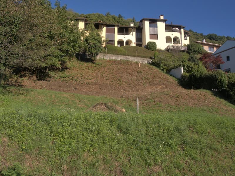 Yellow residential building situated on a slope, with a hill in the background, surrounded by grass and shrubs.