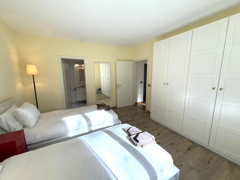 Two single beds, large white wardrobes, wooden floors, a nightstand, a lamp, and a mirror in a hallway