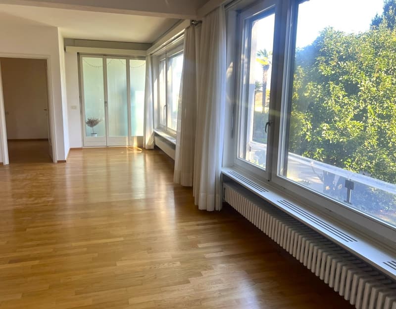 Empty room with hardwood floor, large windows, and curtains. Adjacent room has a sliding door and glass window.