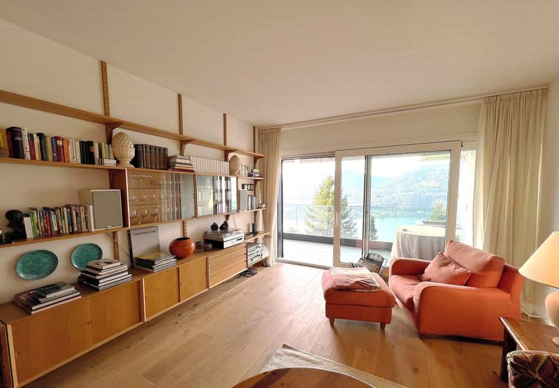 living room with wooden floor, large windows, balcony, couch, ottoman, shelves, books, lamp, table
