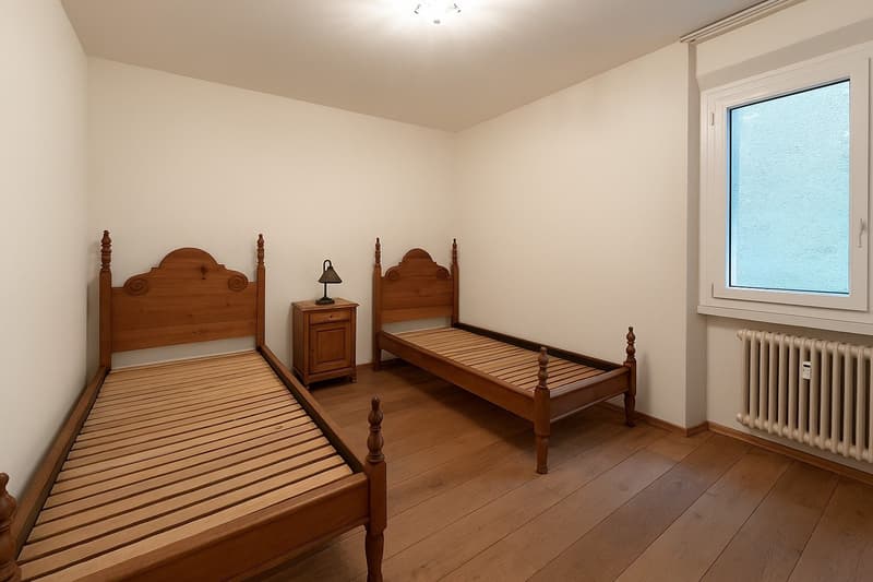 Two wooden beds, nightstand with lamp, white walls, wooden floor, window, radiator