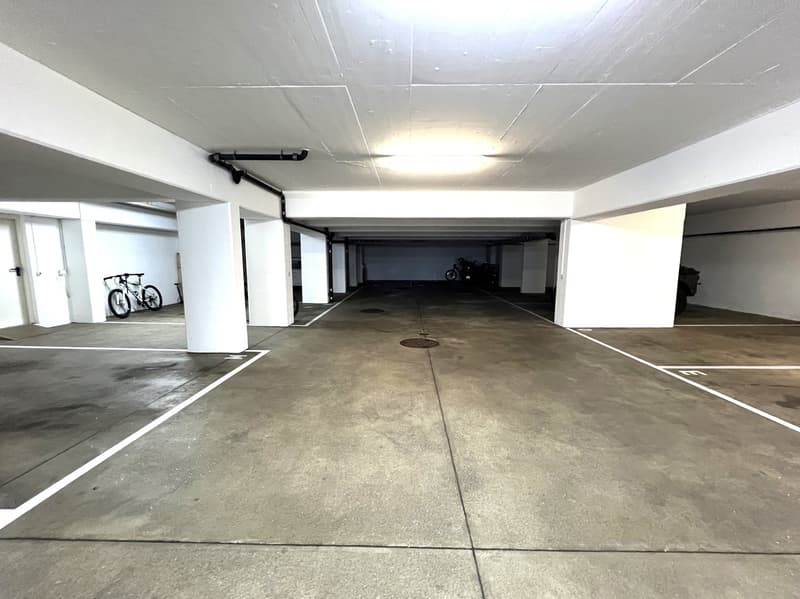 Empty parking lot with white walls and ceiling, concrete floor, two bicycles parked on the left side, a motorcycle parked on the right side, a drain in the middle, and a light on the ceiling.