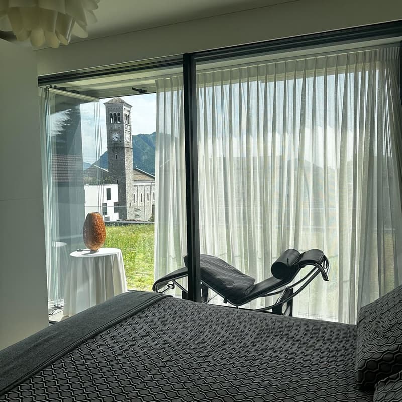 Spacious bedroom with large windows offering a scenic view of a church tower and mountains in the background. The room features a black chaise lounge, a side table with a decorative vase, and sheer curtains covering the windows.
