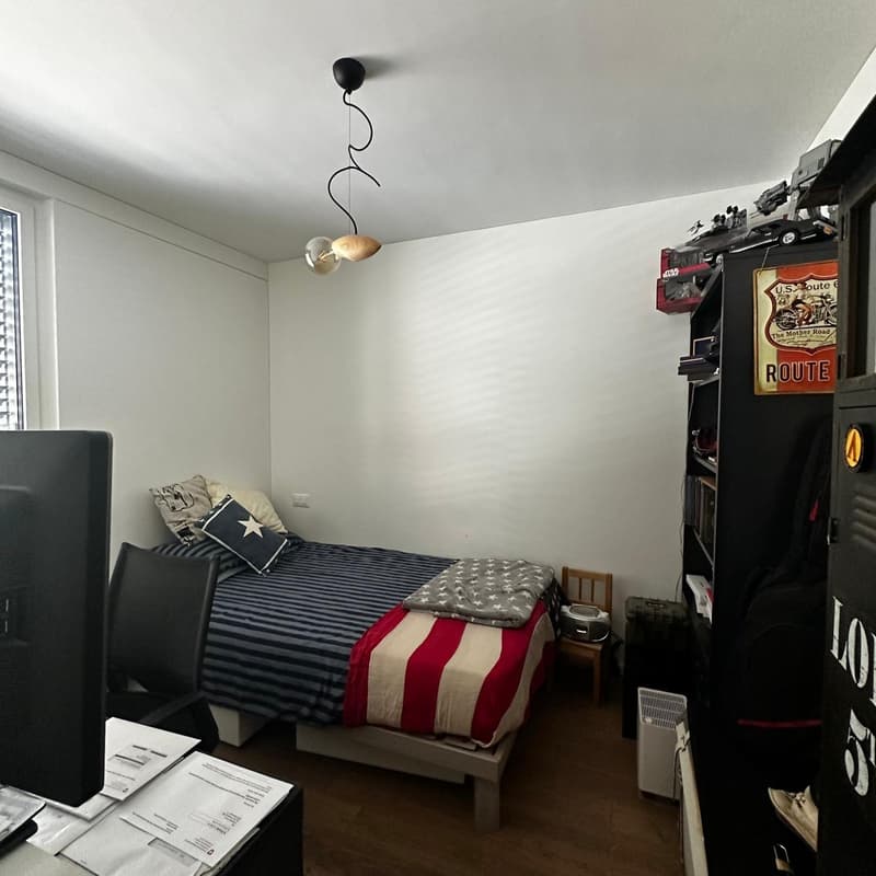 The image shows a bedroom with a bed, bedding with a striped American flag pattern, a desk with papers, and a Route 66 themed wall decor. The room has white walls and a hardwood floor.