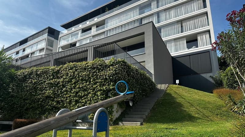 Modern multi-story apartment building with glass balconies, gray exterior, and landscaped grounds with a playground equipment in the foreground.
