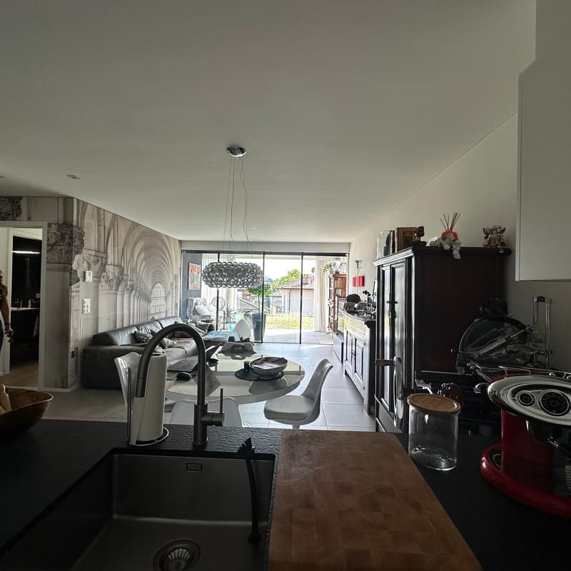Open-concept living room with large windows, modern furniture including a sofa, coffee table, and dining table, kitchen appliances such as a dishwasher, and decorative elements like a chandelier.