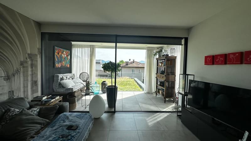 Spacious living room with large sliding glass doors leading to a balcony or terrace, hardwood floors, modern furniture including a sofa and armchair, decorative artwork on the walls, and a shelving unit with various decor items.