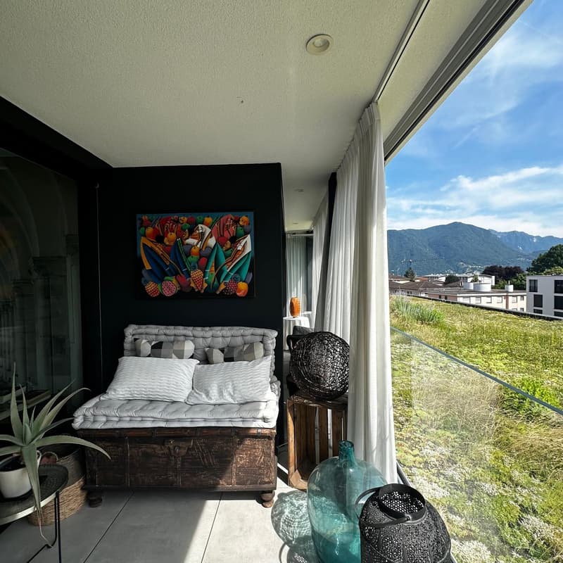 Bedroom with a large window overlooking a scenic outdoor view, a rustic wooden chest serving as a bed frame, colorful abstract artwork on the wall, and decorative elements such as a woven chair and glass vase.