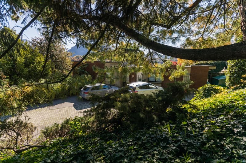 House with brick walls, car parked on driveway, trees, greenery, mountains in the distance
