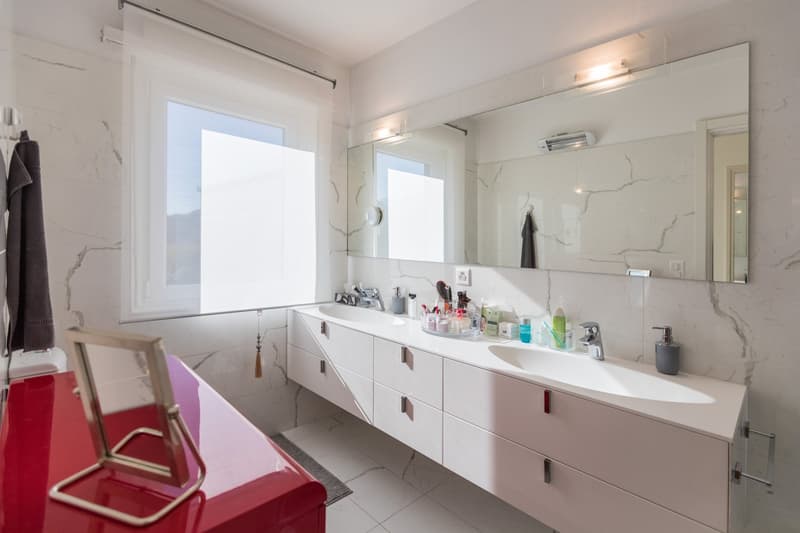 White walls, large mirror, two sinks, red vanity with mirror, power outlet, faucet, window