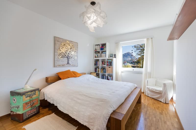 wooden bed, orange pillows, white bedspread, chair by the window, small cabinet with lamp, book shelves, painting above bed