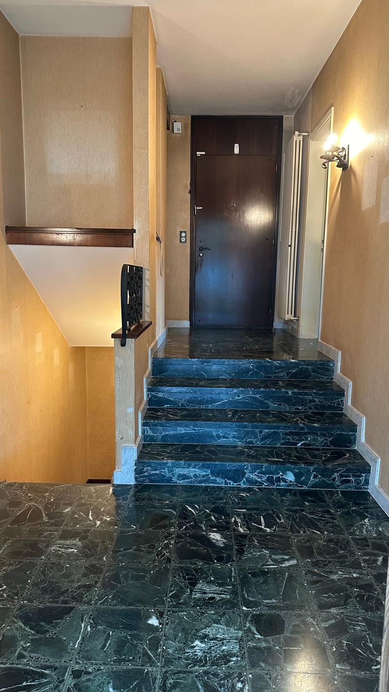 The image shows a hallway with a dark marble staircase leading up to the entrance door. The walls are painted in a light, neutral color, and there is a wooden shelf or ledge along one wall. The entrance door appears to be a dark, wooden door.