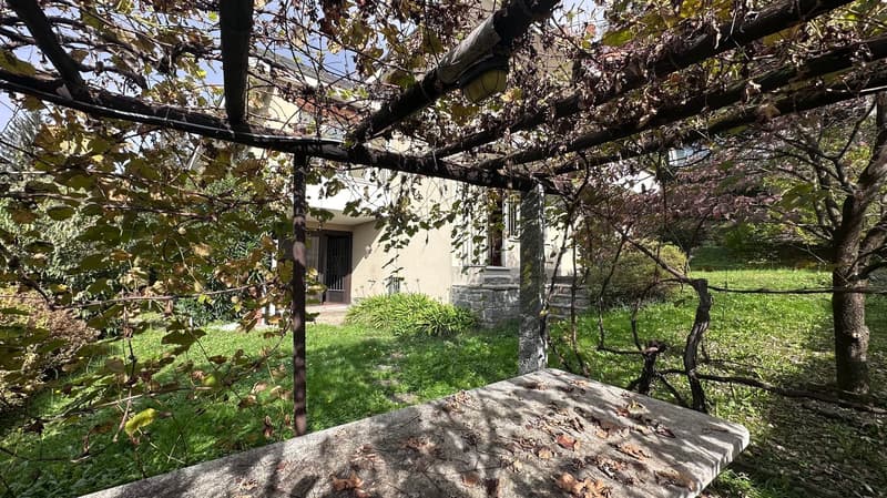 A stone house surrounded by lush greenery, including a grassy lawn and trees with autumn foliage. There appears to be a balcony or terrace visible on the side of the house.