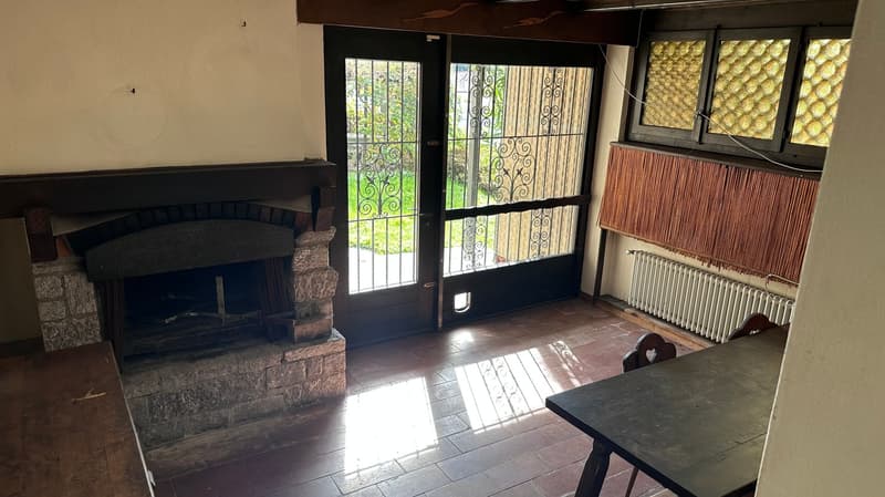The image shows a cozy living room with a stone fireplace, large windows with decorative wrought-iron grilles, and a wooden table. The room has a rustic, traditional feel with the exposed wooden beams and tile flooring.