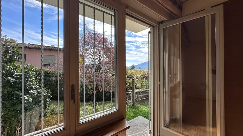 Large windows with a view of a garden and mountains, wooden framing, stone floor, and a balcony or terrace visible through the open door