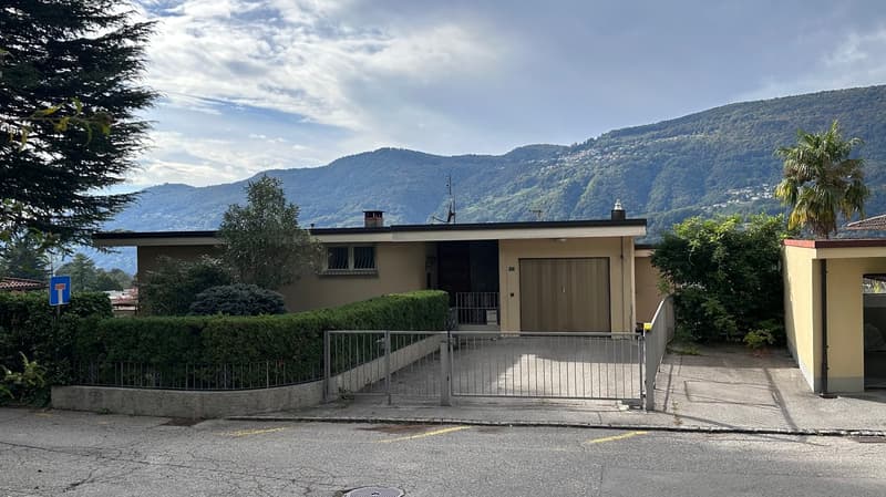 Single-story house with a garage, surrounded by trees and mountains in the background. The house has a beige exterior and a metal gate at the entrance.