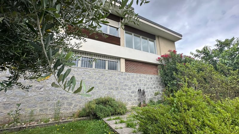 2-story house with stone exterior, large windows, balcony, and surrounded by lush greenery including trees and shrubs