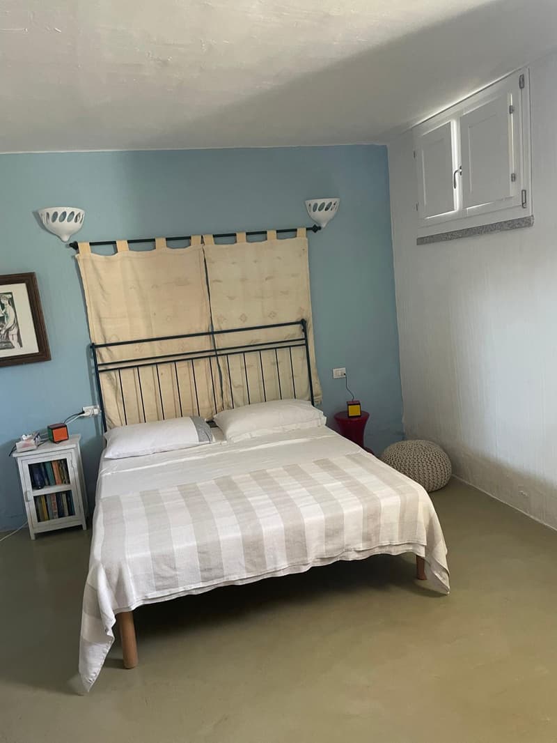double bed with white pillows, nightstand with books and charger, red stool, beige walls, white ceiling, lamps, curtain, power outlet