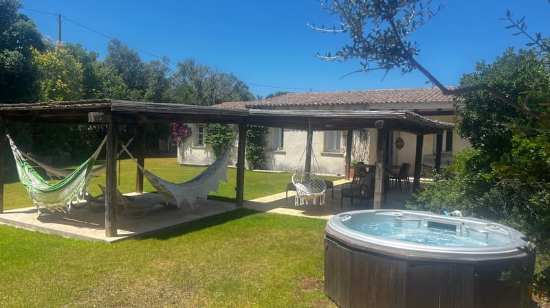 grass, wooden pergola, 2 hammocks, round hot tub, green plants, trees, patio with table and chairs