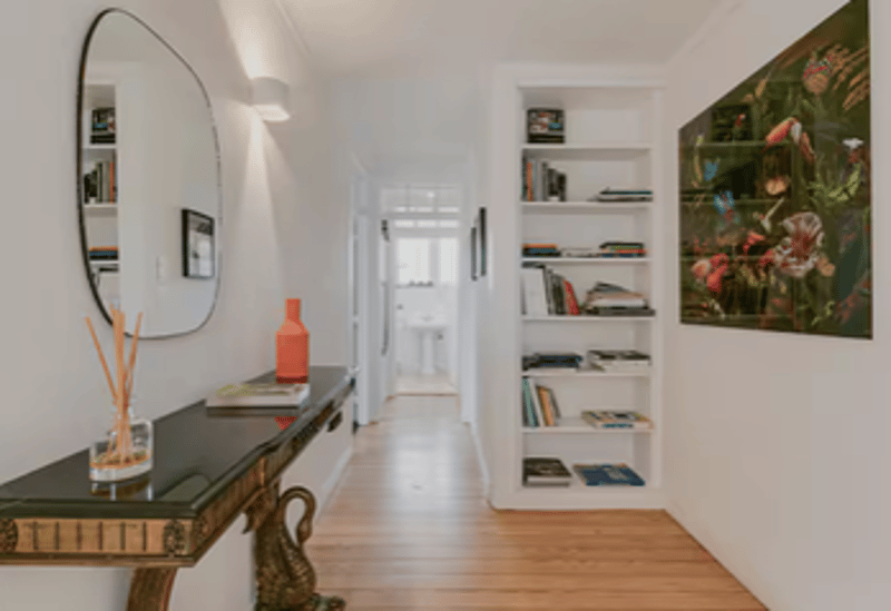 A hallway with wooden floor, white walls, a mirror, a table with a vase, a shelf with books, and a painting on the wall.
