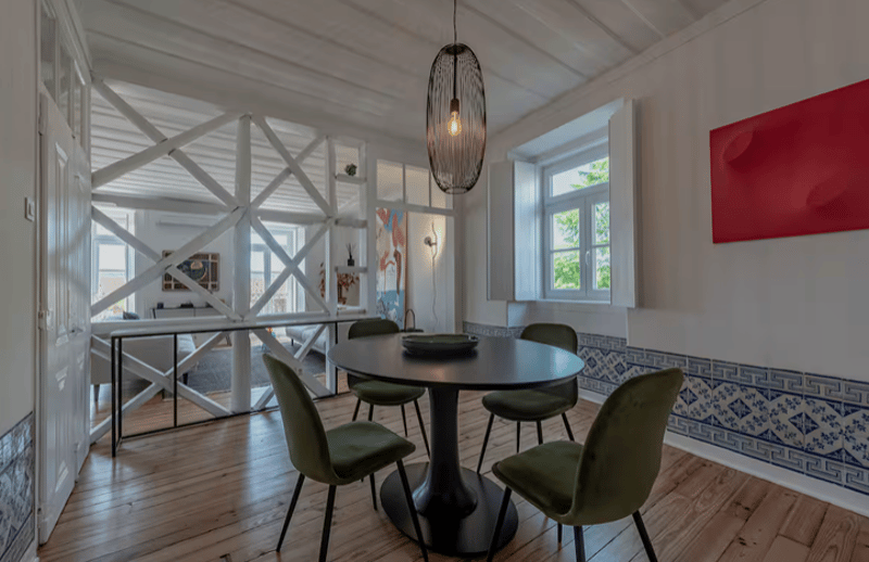 Dining room with wooden floor, round table, green chairs, white walls, blue and white tiles, hanging lamp, windows, metal railing