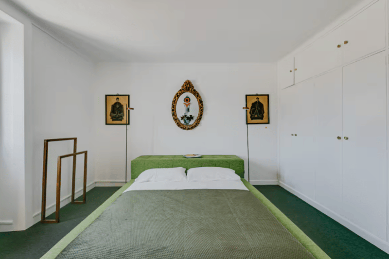white walls, green carpet, bed with pillows and blanket, artwork on the wall, lamps