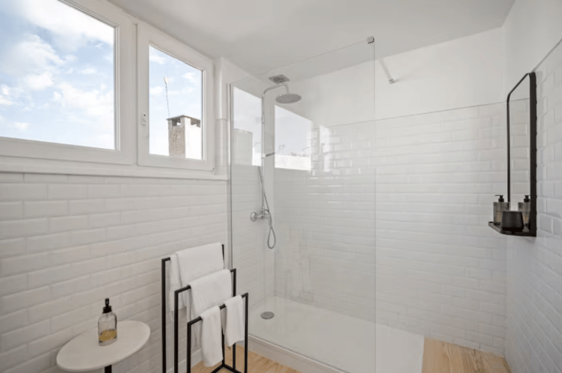 shower stall, white tiled walls, glass door, shower head, towel rack, table with a bottle