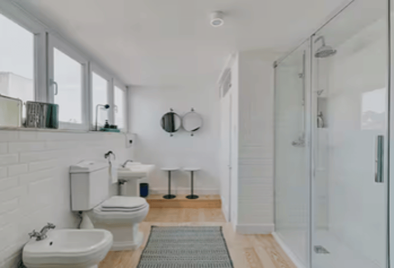 white washbasin, white bidet, toilet, glass shower enclosure, white tiles, large windows, wooden floor