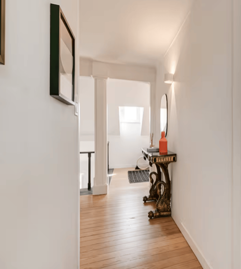 hallway with wooden floors, white walls, mounted frames, decorative mirror, ornate console table, lamp