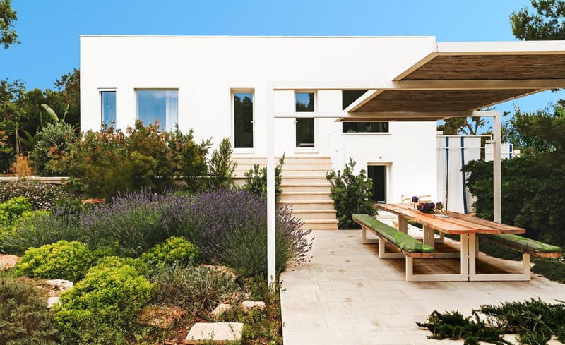 Modern white house, large windows, wooden pergola, outdoor dining area, garden with various plants
